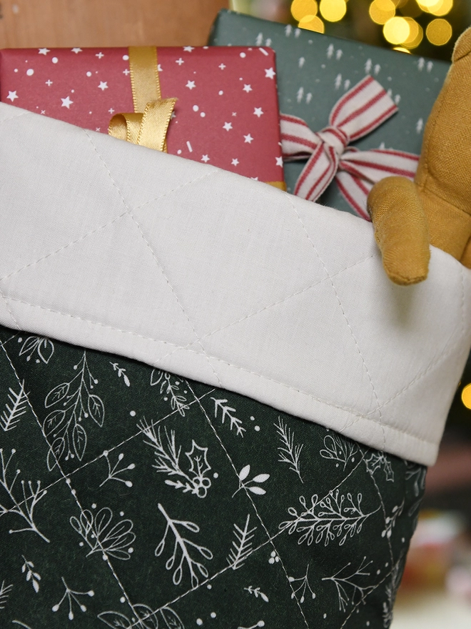 A patchwork stocking in green colours with an ivory turnover cuff hangs on a wooden door. Small gifts and toys poke out of the top.