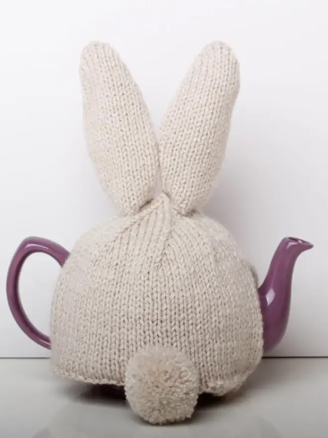 Hand Knitted Bunny Rabbit Tea Cosy, a rabbit bunny tea cosy sitting on a grey surface against a white backdrop. It fits over a purple tea pot. 