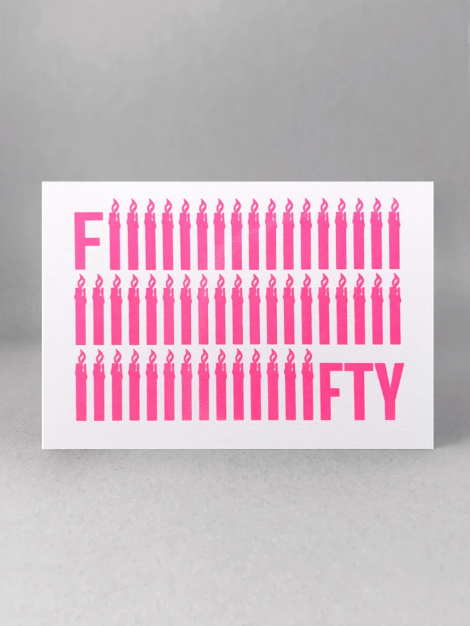 The word Fifty screen printed in neon pink ink across a landscape rectangular recycled white card. To emphasise the fifty, the letter i is shaped like a simple lit candle and is repeated fifty times in three rows. The card is stood square on in a light grey studio set 