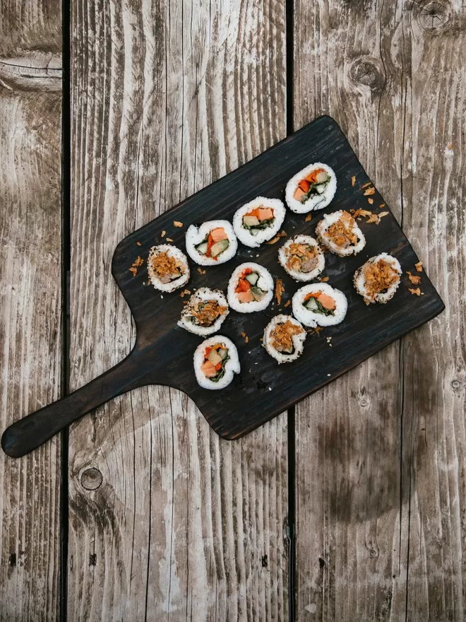 ‘Fancy A Snack?’ Long Handle Charred Black Serving Board seen on a wooden table with sushi on top.