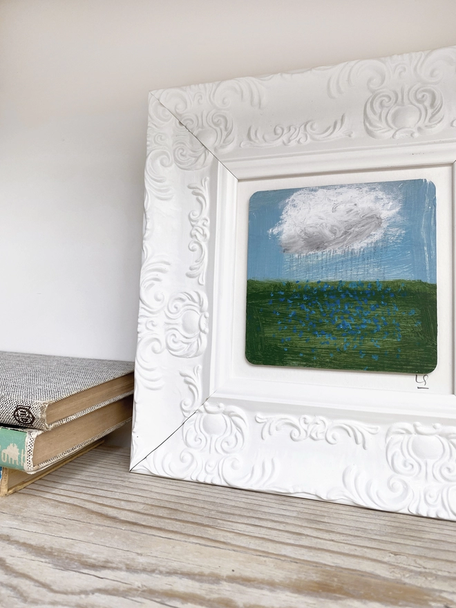 wooden shelf with painting of blue sky, a cloud and a field of blue forget-me-nots in chunky white ornate frame