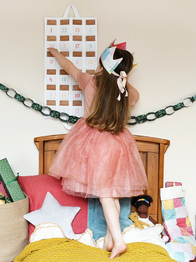 A young girl wearing a pink tutu dress stands on a bed facing an advent calendar hung on the wall. It is made of fabric with 24 pockets each containing a wooden story card.