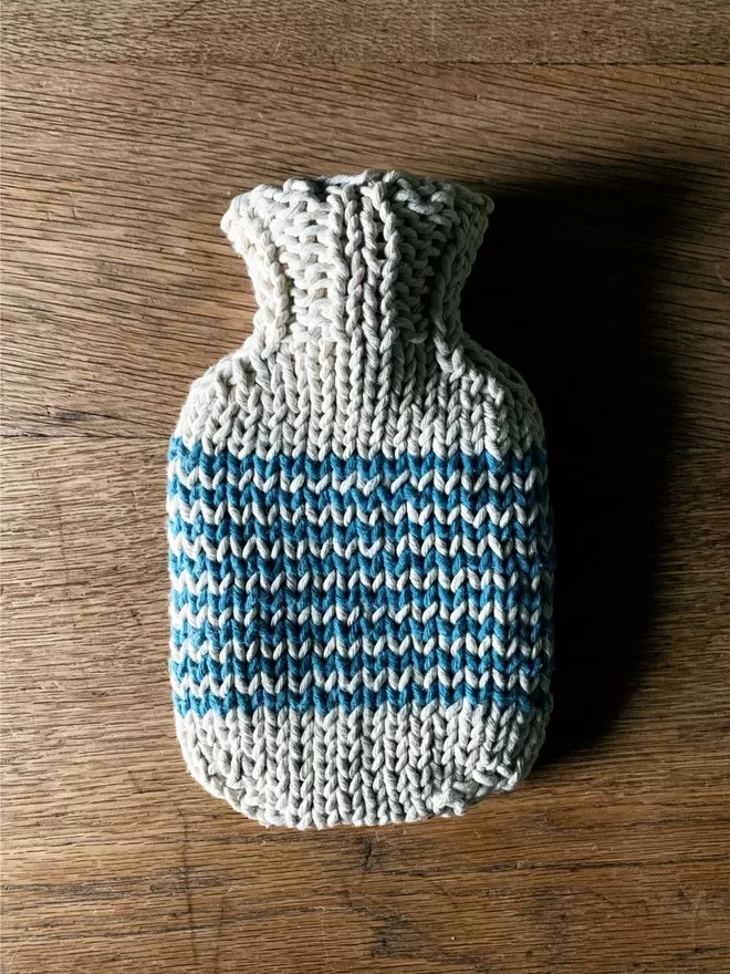 Mini Hot Water Bottle in hand knit string cover with blue stripe on an oak table