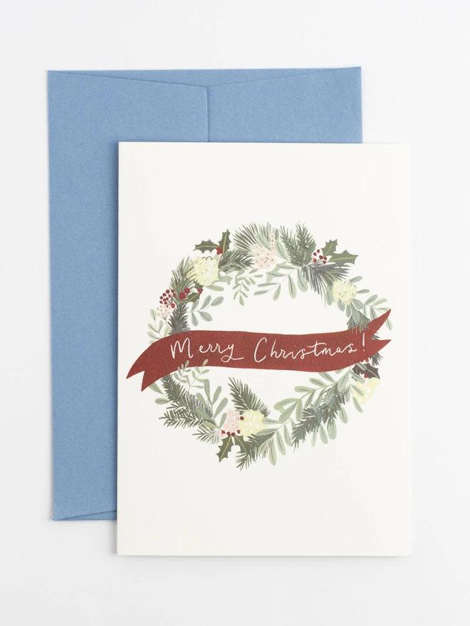 A Christmas card with an illustrated christmas wreath design of green foliage and pink and red berries. Across the centre of the wreath is a ribbon with lettering that reads "Merry Christmas!"