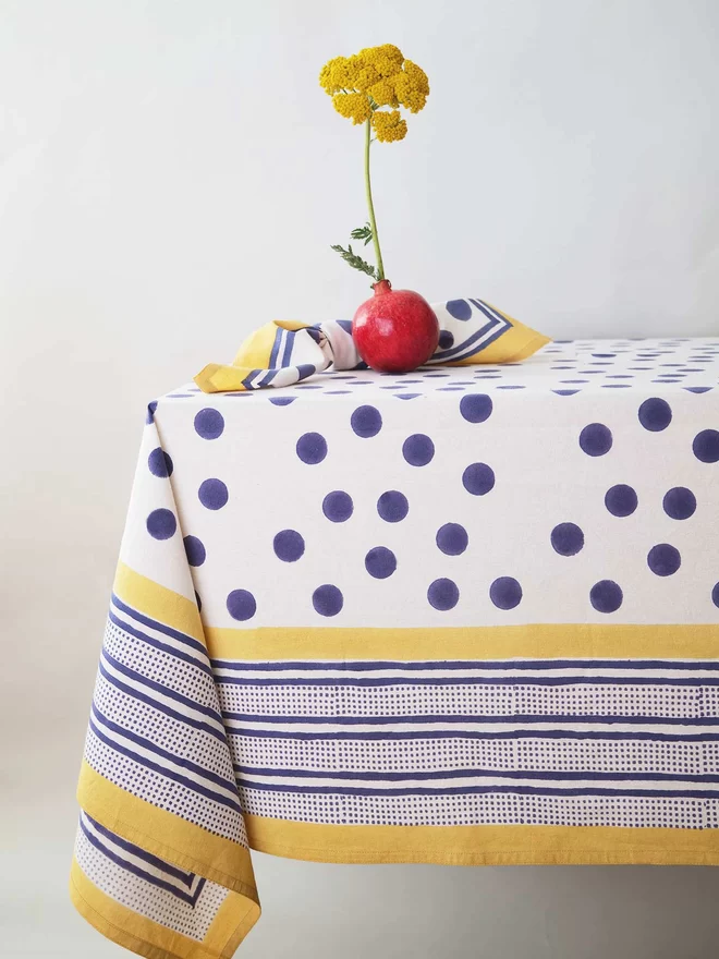 Block printed cream tablecloth featuring a navy spot and yellow border paid on a table with matching napkin and a pomegranate used as a vase and holding a yellow flower