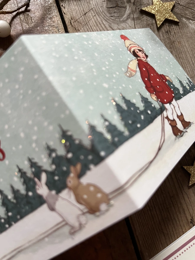 a close up of a Christmas greeting card featuring a vintage style illustation of a little girl with a brown bob pulling a sledge finished with glitter glue