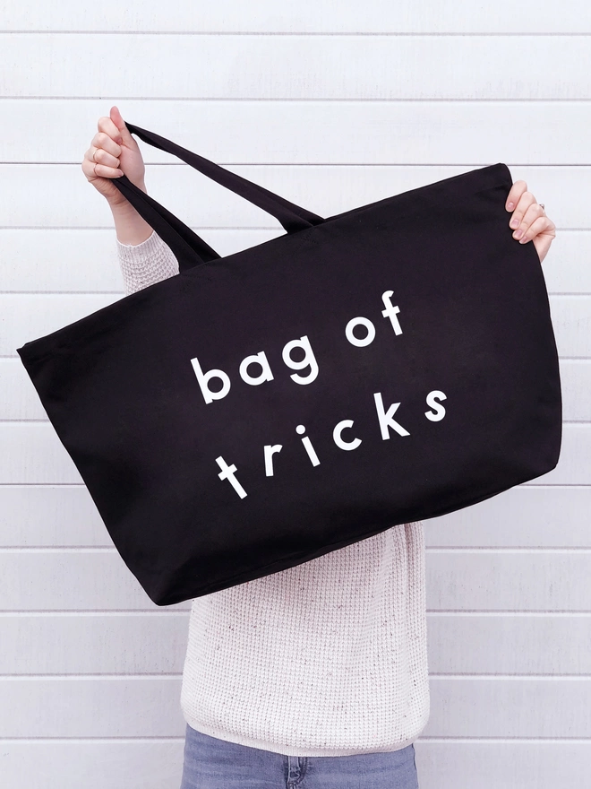 model holding out an oversized black canvas tote bag with the words bag of tricks