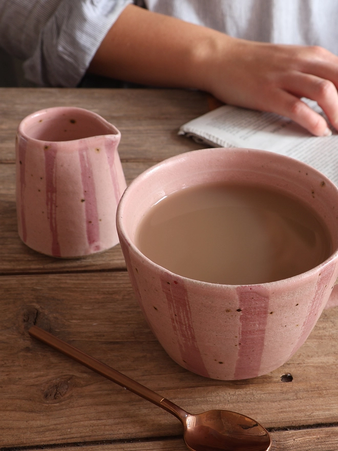 Handmade Ceramic Stoneware Mug in Pink Stripe sat on a wooden surface. The mug is filled with coffee and is surrounded by a brass spoon, a small jug in the same pink stripe design and a newspaper with a female arm resting on top. 