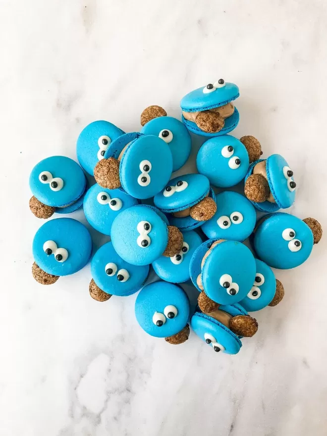 blue cookie monster macarons with googly eyes and a chocolate chip cookie in its mouth, sitting in a pile on top of each other