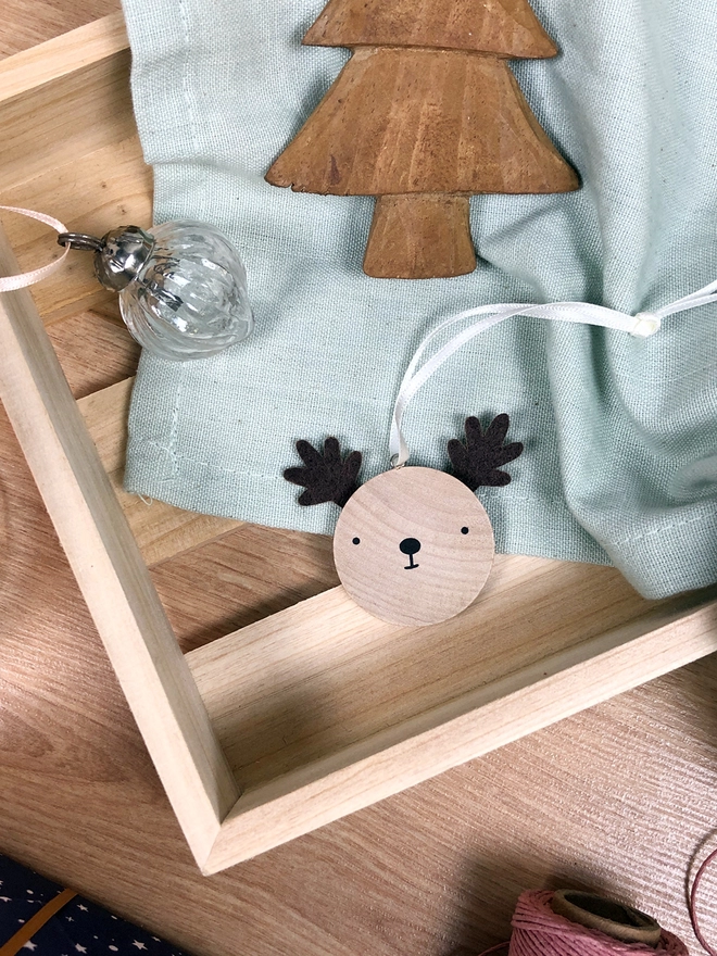 A small wooden and felt reindeer decoration lays in a wooden crate alongside other Christmas decorations.