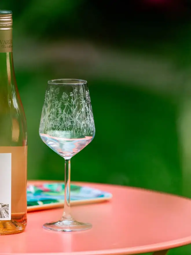 Meadow Wine Glass, an etched wine glass with a wildflower pattern sits on a bright pink patio table next to a bottle of rosé wine.