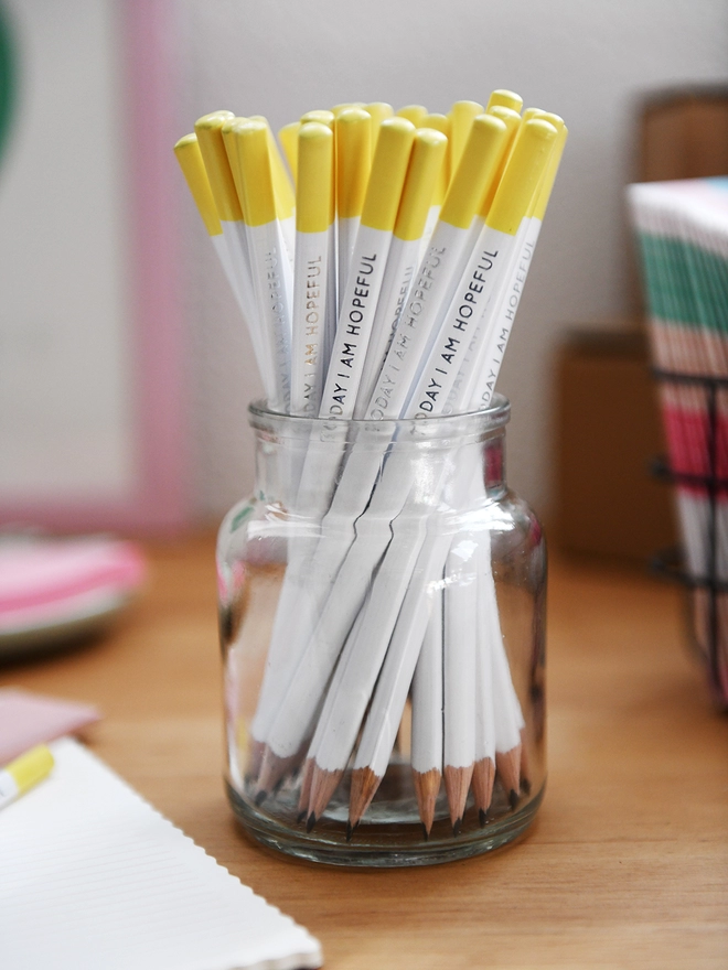 A glass jar full of white pencils with yellow tips stands on a wooden desk surrounded by various stationery items.