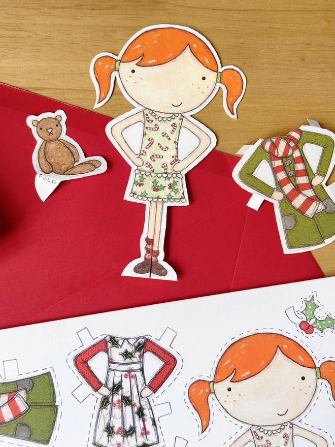A Christmas card with an illustrated paper doll design is with a red envelope on a wooden desk.