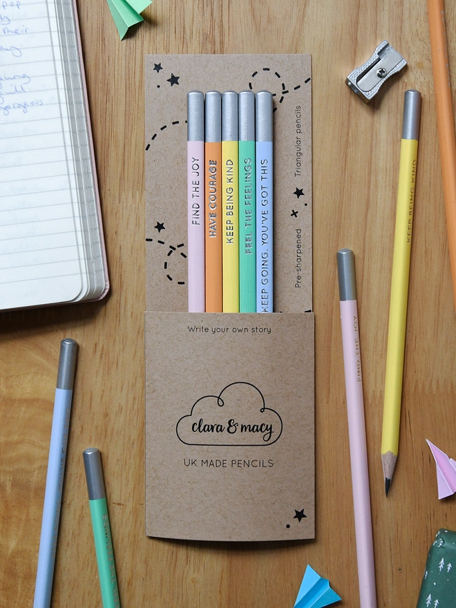 Five pastel pencils in cardboard packaging lay on a wooden desk, with four pencils and a notebook around them.