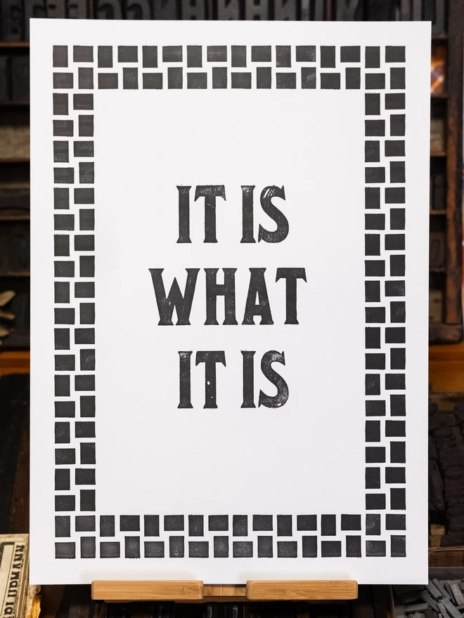 A3 size letterpress print with vintage serif block font text design "It is what it is" bordered by vintage woven squares design