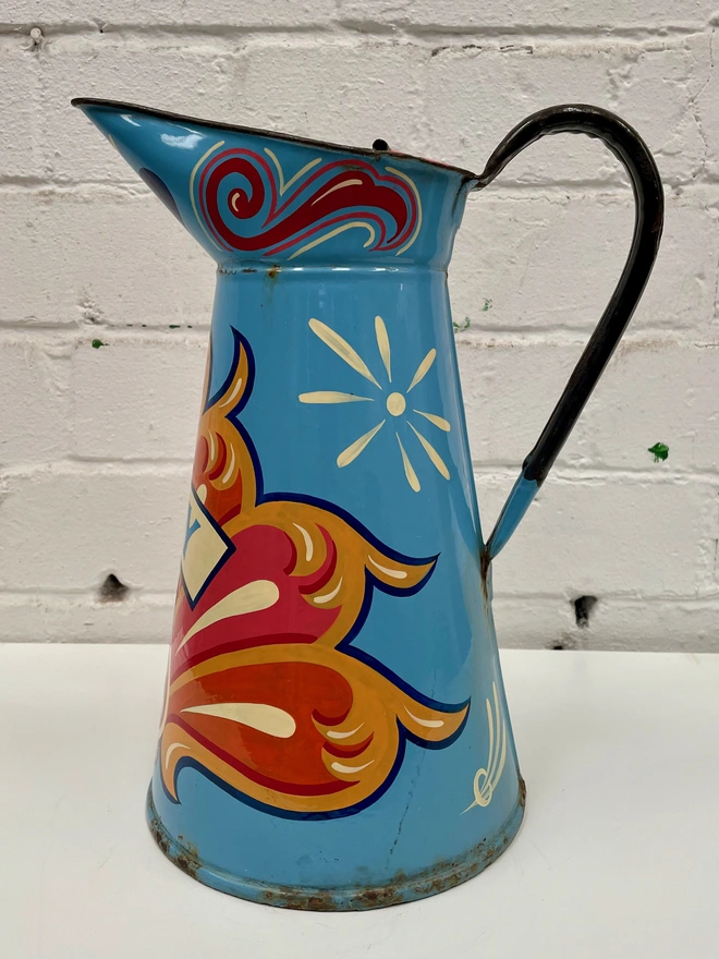 Large blue enamel jug hand-painted with a traditional fairground inspired design, in pink, orange and yellow. 'Joy' is painted in a banner across the centre.