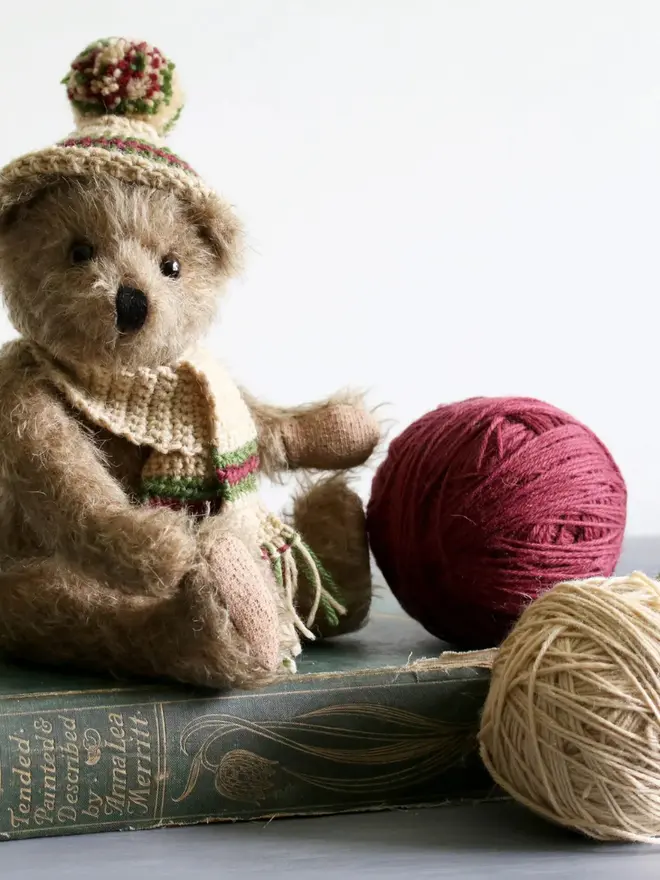 Brown teddy bear sitting upright against a white background among round balls of wool, wearing a knitted beige hat and scarf with green and burgundy details.