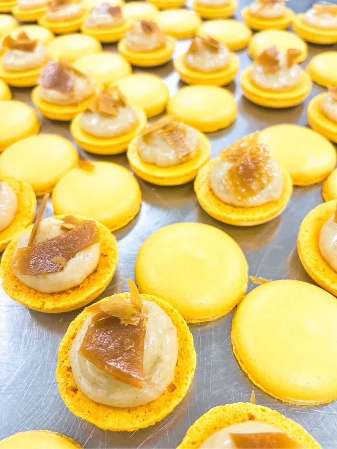 rows of yellow macarons showing their filling of stem ginger white chocolate ganache with a buttery caramel crisp toping