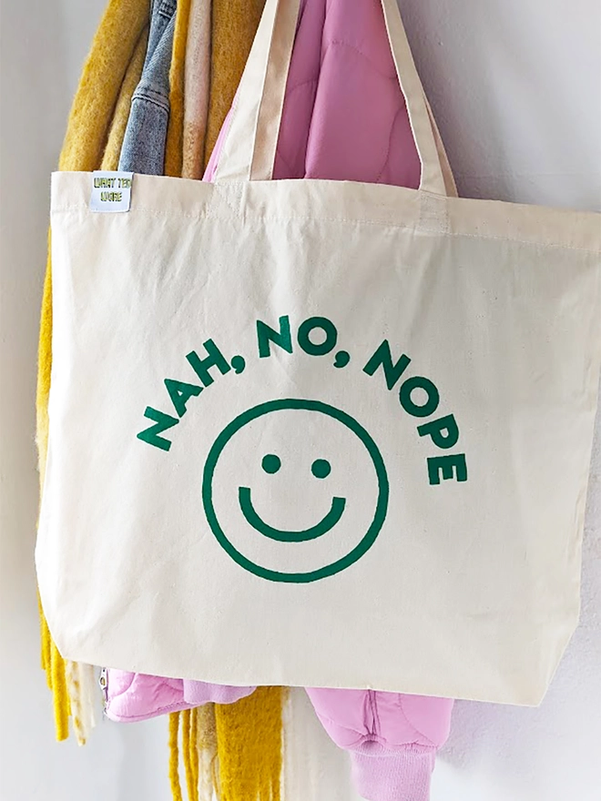 A hanging natural canvas tote bag with green Nah, No, Nope slogan and a graphic smiley face.