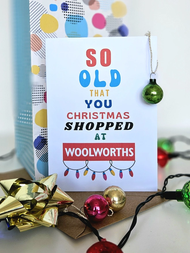 A photograph of the front of a humorous A6 greeting card illustrated using brightly coloured text and the retro shop logo. The text says so old you Christmas shopped at Woolworths with garland lights