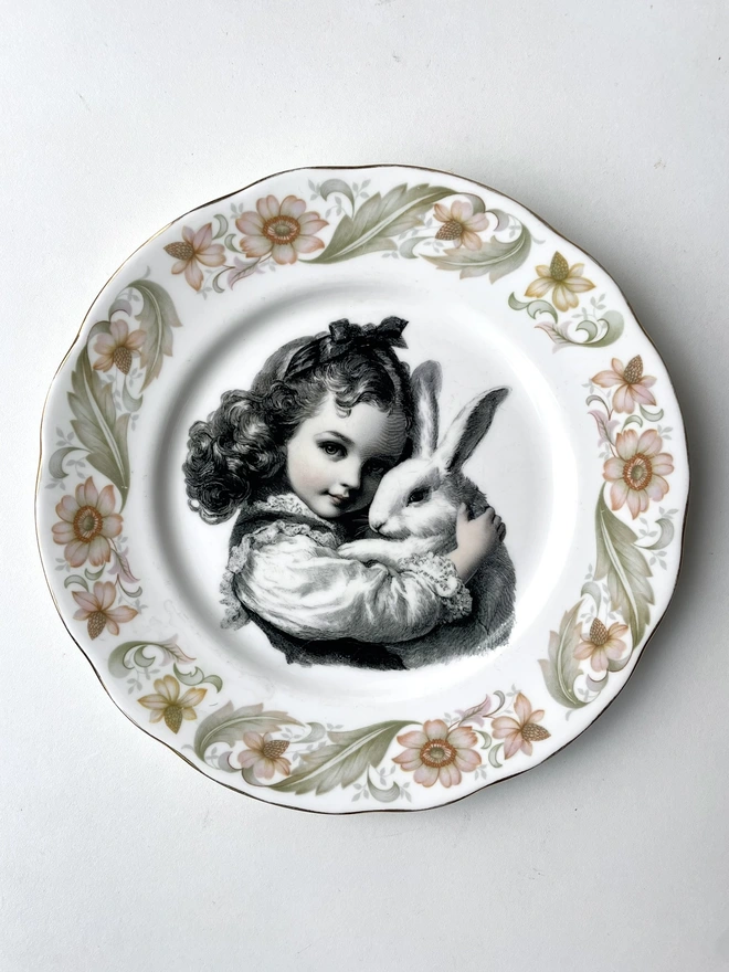 a vintage plate with decorative ornate borders, in the centre i have printed a vintage black and white illustration of a little girl holding her pet white bunny rabbit