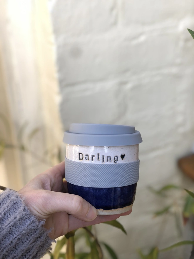 Personalised Short Ceramic Travel Mug, person holding their blue mug in their hand indoors in front of a white brick wall. Each mug has their own unique personalisation. 