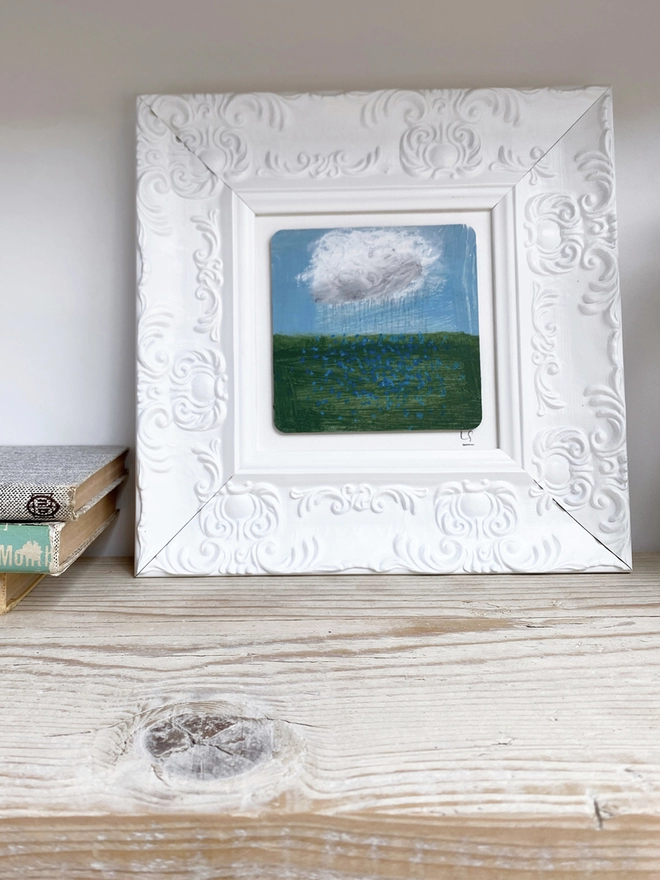 wooden shelf with painting of blue sky, a cloud and a field of blue forget-me-nots in chunky white ornate frame