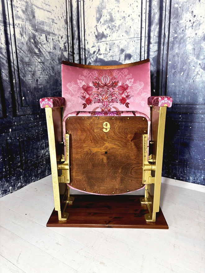 A vintage cinema seat fully refurbished with pink patterned velvet upholstery, the seat is mounted to a wooden plinth and is sat on a white wooden floor against a blue marbled background