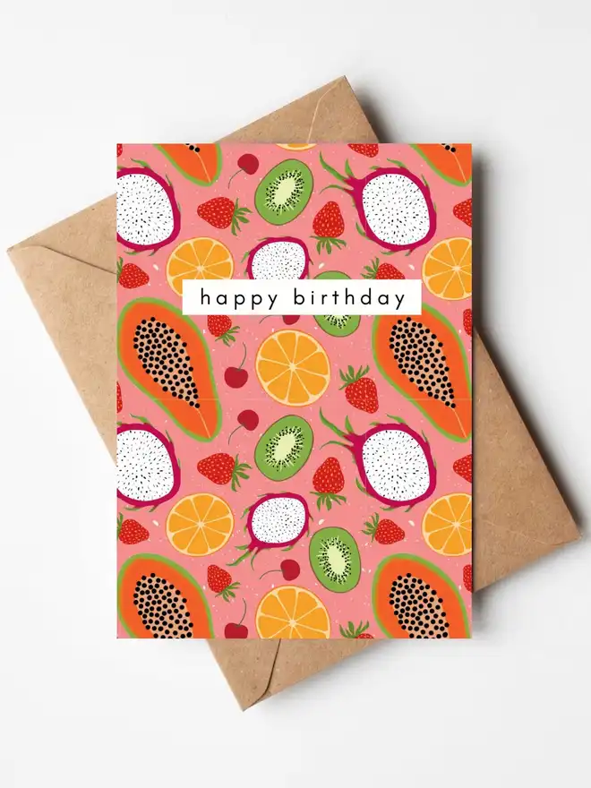 A colourful birthday card featuring hand-illustrated tropical fruits including papaya, dragon fruit, kiwi, oranges, strawberries and cherries on a pink background, with the words "happy birthday" across the centre. The card is displayed with a kraft envelope.