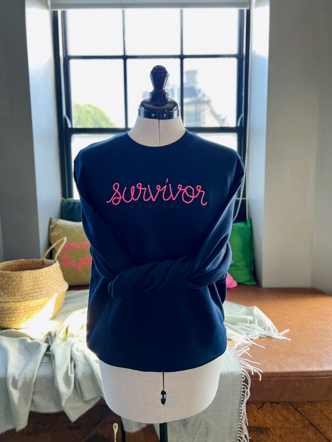 A navy sweatshirt on a tailors dummy embroidered with survivor in pink 