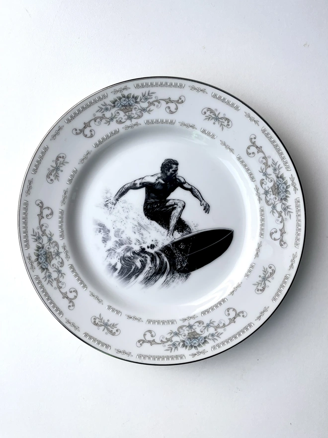 a vintage style image of a surfer riding a wave, 60s style, is printed in the centre of a vintage plate with an ornate edge