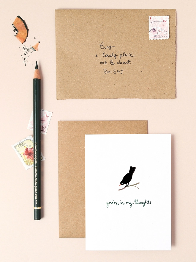 An image of a mini, rectangular white greetings card with an illustration of a black bird perched on a small branch with a hand written 'you're in my thoughts' message in black ink seen under the central illustration. The card is placed on top of a brown rectangular kraft envelope.