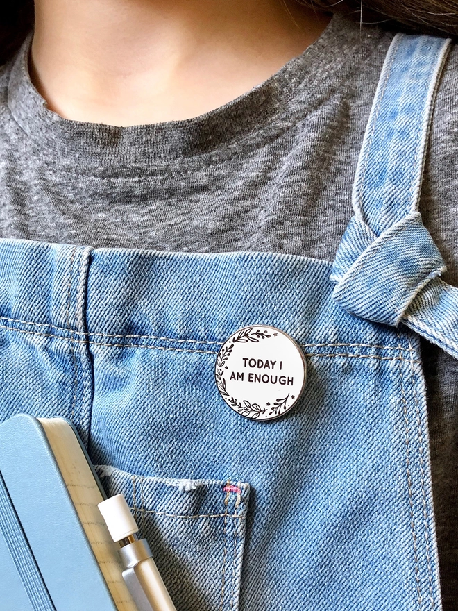 A white enamel pin is pinned to someone's blue dungarees. The pin is round with a botanical design and the words "Today I Am Enough".