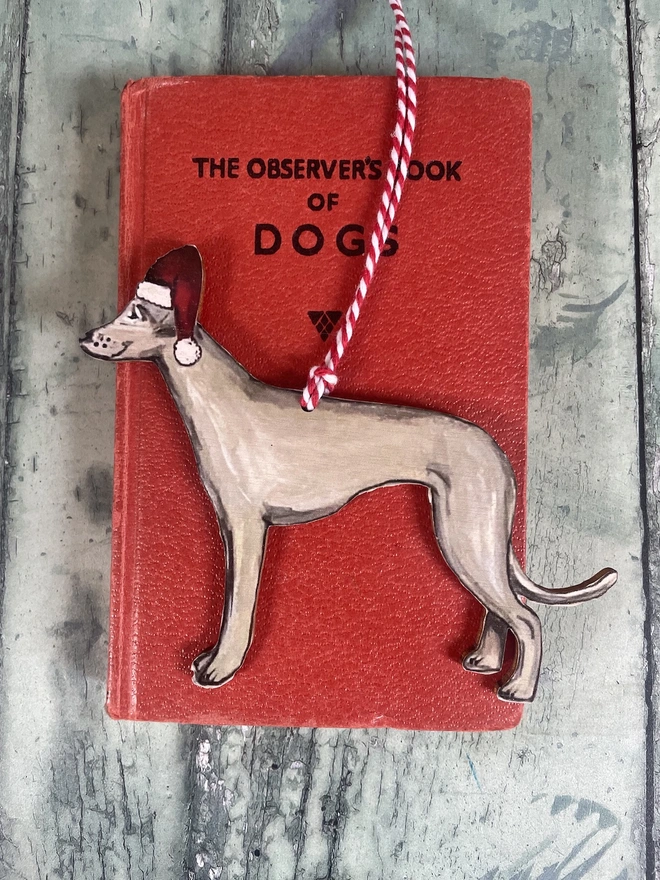 Whippet wooden Christmas decoration wearing a santa hat hung with red and white twine placed onto a red book about dogs