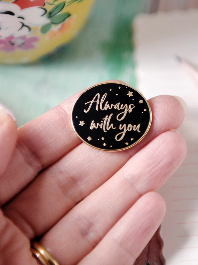 A black and gold enamel pin badge rests on a hand. It has a subtle star design and the words "Always with you".