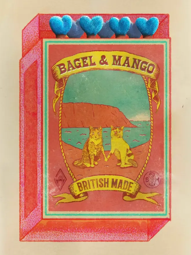 
An illustrated, retro-style matchbox with a bright red and orange border. The central image shows two cats, one a pale ginger and the other a light yellow, sitting side-by-side on a beach. Behind them is a large red cliff face and a body of water with a distinct shoreline. The scene is framed by a knotted rope border. A banner at the top reads "BAGEL & MANGO," and another banner at the bottom says "BRITISH MADE." The match tips on the top are heart-shaped and a vibrant blue. The entire image has a grainy, aged texture.