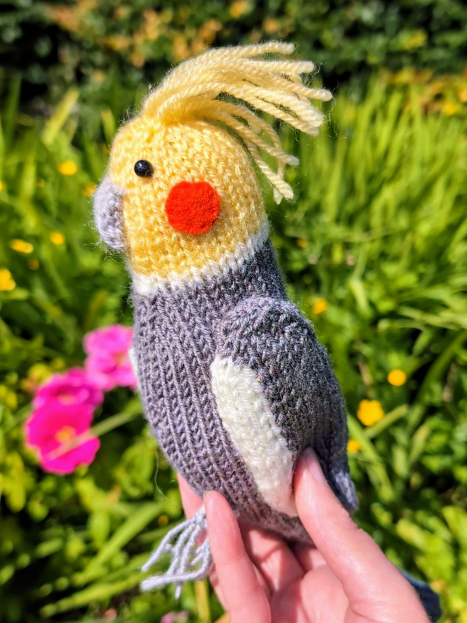 a knitted cockatiel being held in a garden of colourful flowers