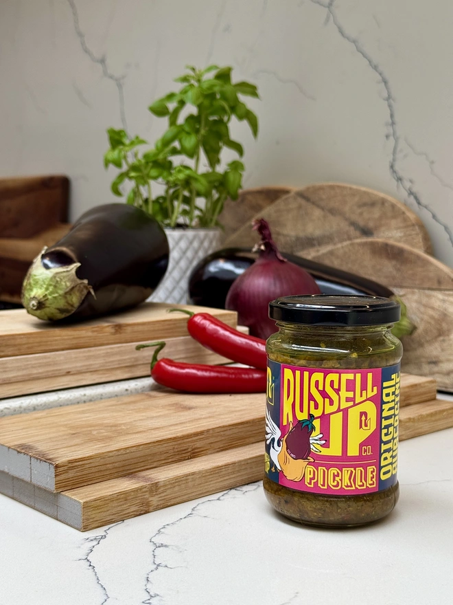 father's day pickles - cheese set. a jar of aubergine pickle photographed next to a chopping board and onion, aubergine and chilli.