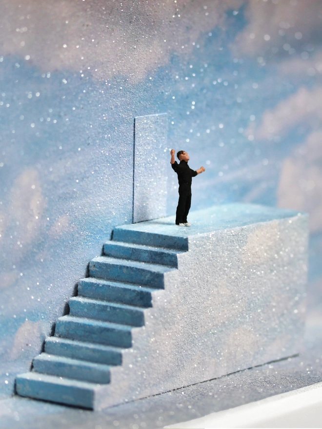 Diorama in which miniature Truman stands at the top of the stairs, ready to open the door to reality., set against a sparkling sky