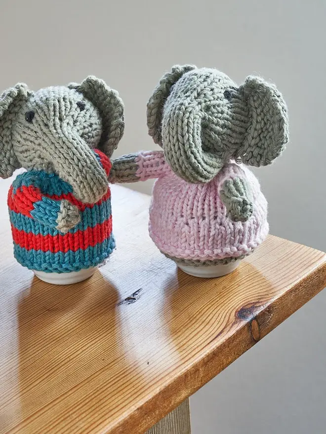 Egg Cosy Elephant Table Decoration (Set Of 2), a pair of colourful elephant cosies sitting on a wooden surface. 