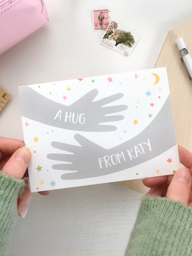 A friendship greetings card with a hugging arms design and personalised wording that reads "A hug from..." is being held above a white desk.