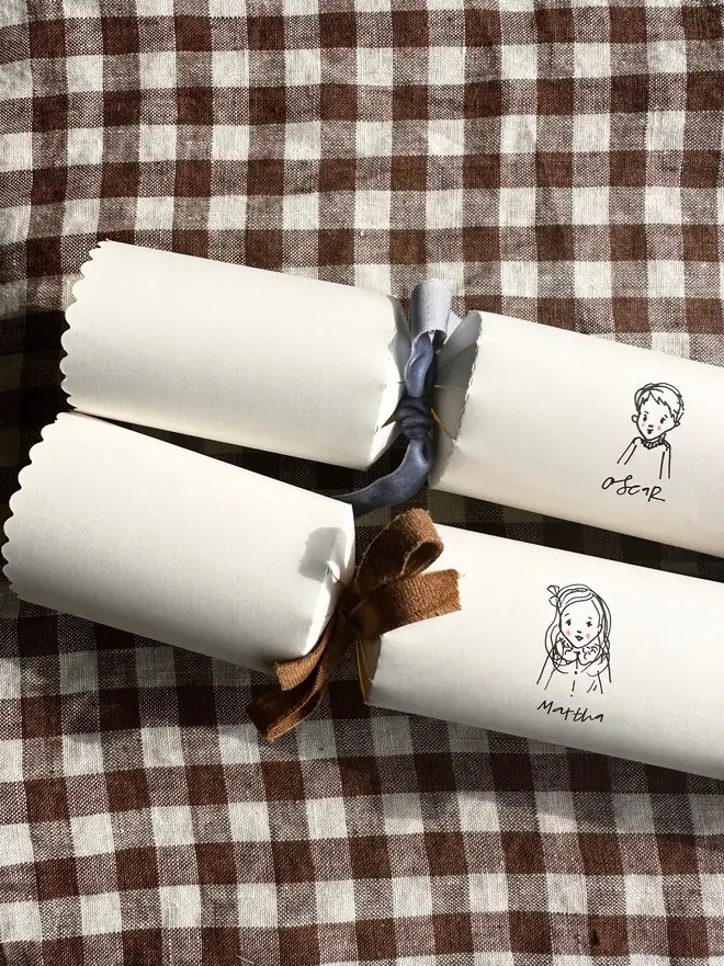 two white christmas crackers, each with a bespoke illustration of a child or pet, with different coloured velvet ribbon. sitting on brown and white gingham table cloth