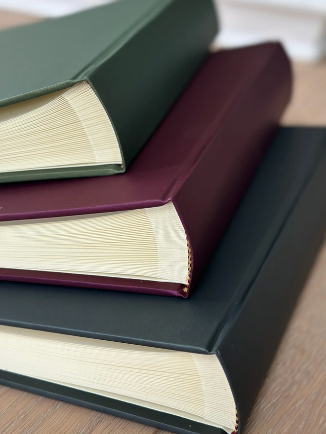 A pile of hardback photo albums rests on a wooden surface. The colours are green, plum and black.