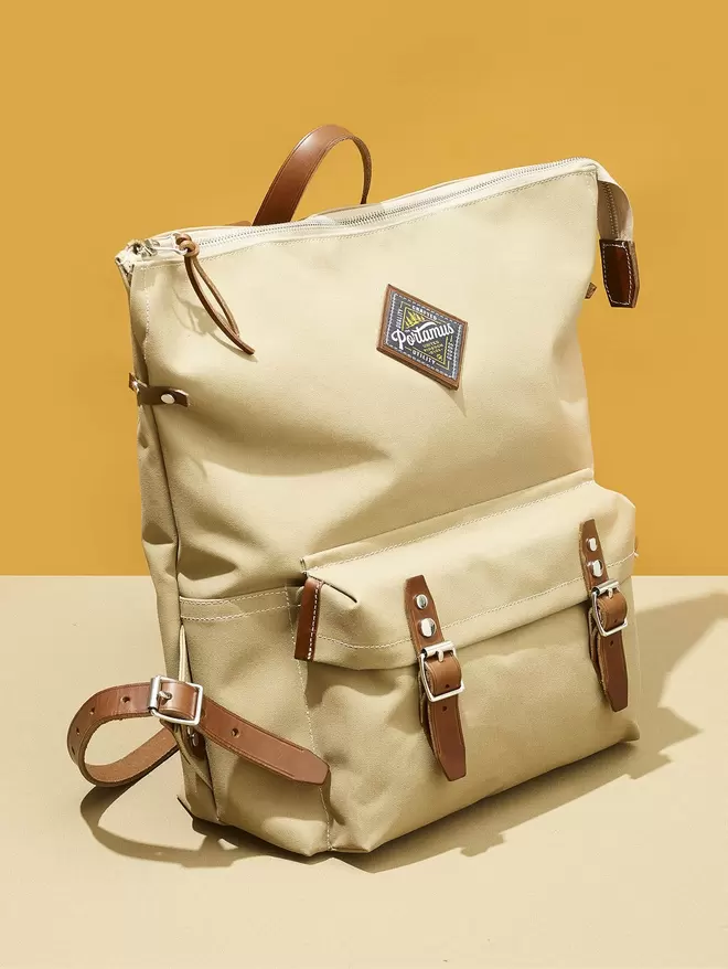The Shortwood zip top back pack in Scout taupe with brown leather straps, on a yellow and taupe two tone backround.