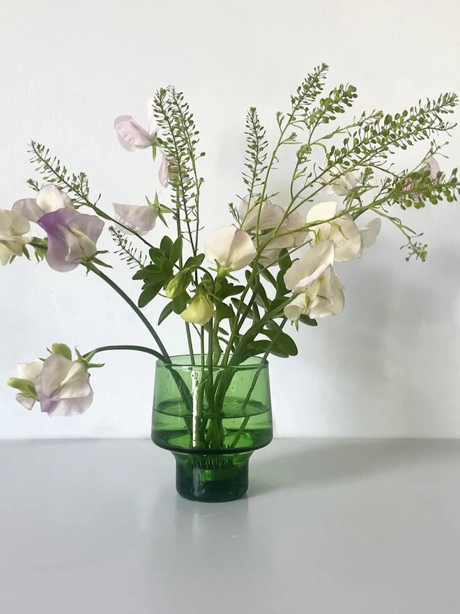 Small Green Flower Arranging Items (Set Of 2), a green glass vase sitting on a plain surface against a white backdrop. It is filled by a bundle of flowers grouped together by a copper frog. 