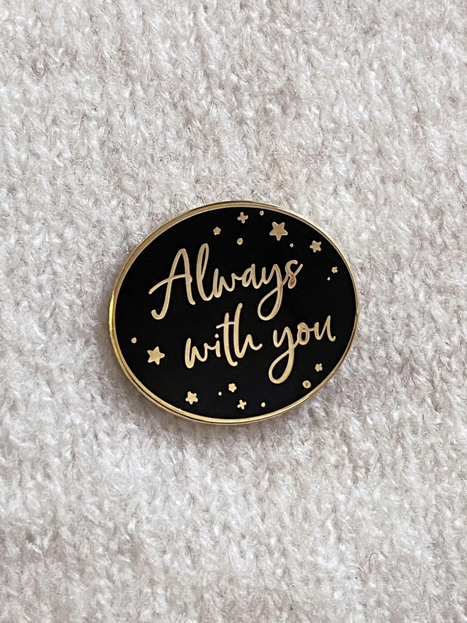 A black and gold enamel pin badge is pinned to a beige jumper. It has a subtle star design and the words "Always with you".
