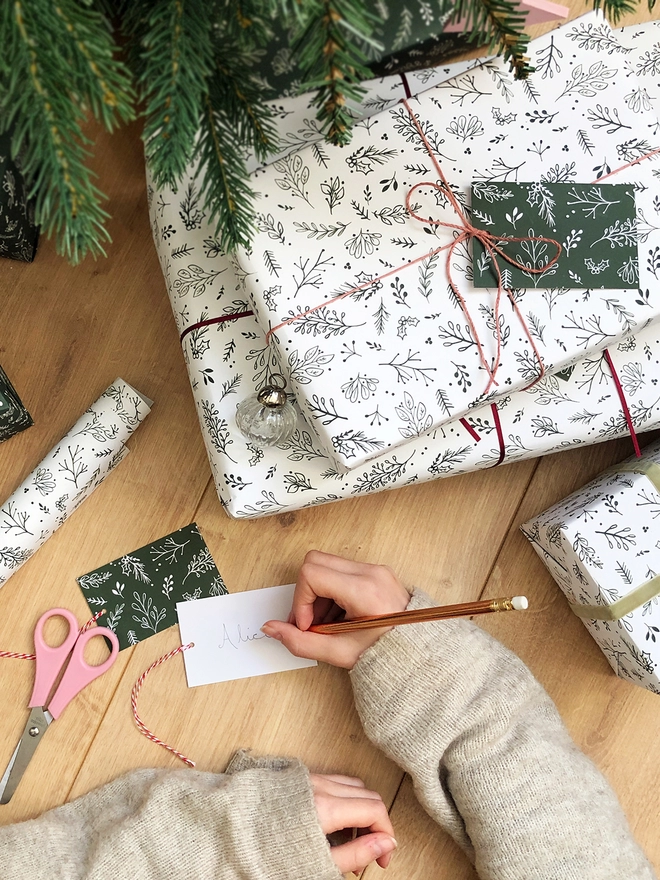 A pile of gifts wrapped in white wrapping paper with a green foliage design lay on a wooden floor beneath a Christmas tree.