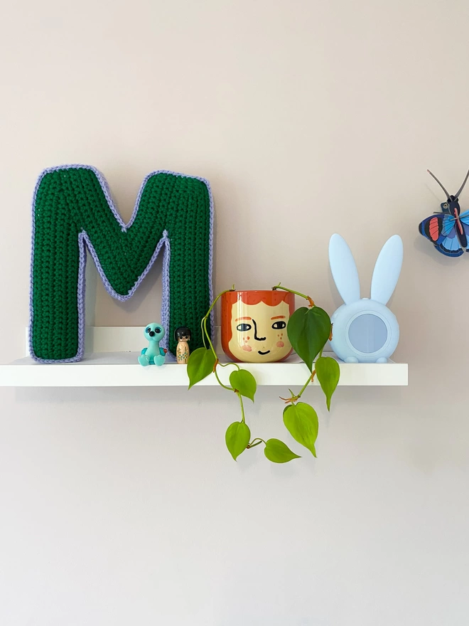 Cushion shaped like the letter M in Grass Green and Lilac, on a child's shelf