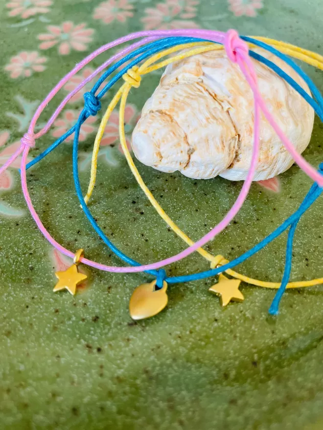 Three wish bracelets with gold charms resting on a shell on a green plate.