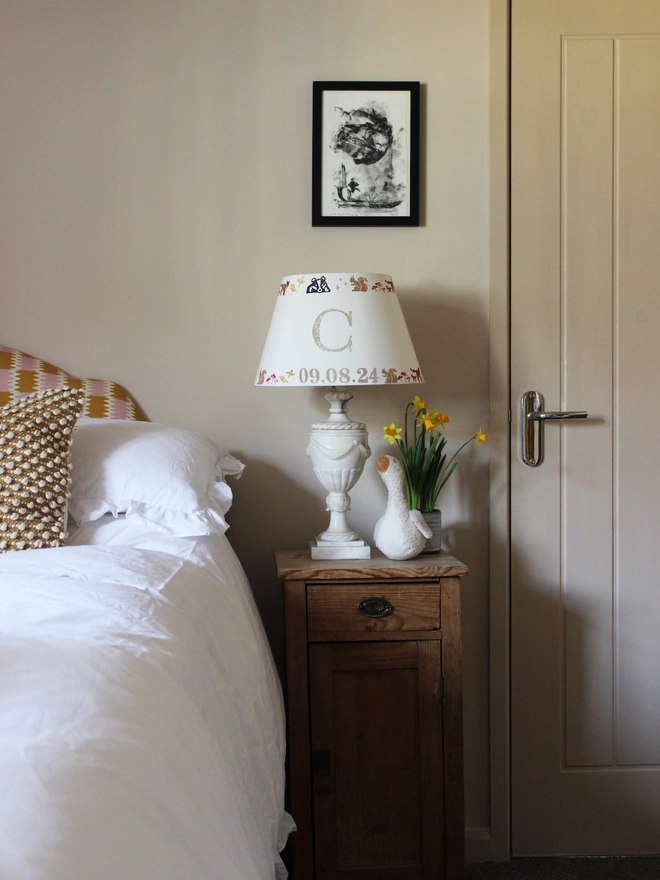 A cosy children's bedroom with a rustic wooden nightstand featuring a personalised lampshade with the letter "C" and a countryside pattern. A plush goose, daffodils, and white bedding add charm. Neutral empire light shade with pretty animal details, ideal for kids bedroom design and personalised kids gift.